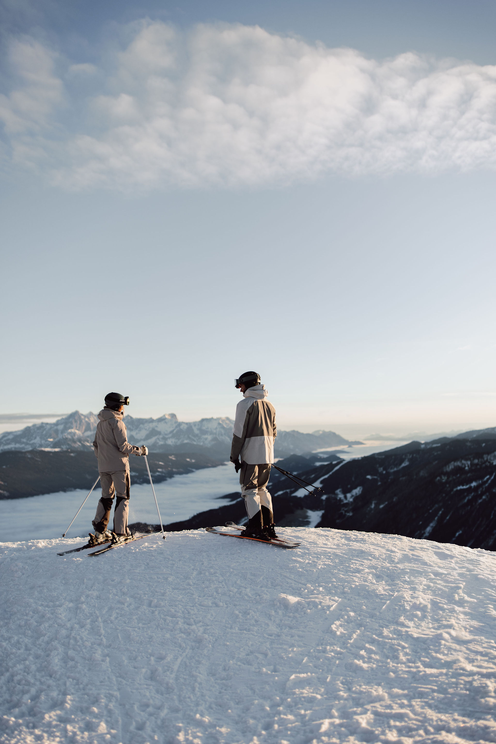 Two skiers stand on a snowy mountain overlooking distant peaks and a lake